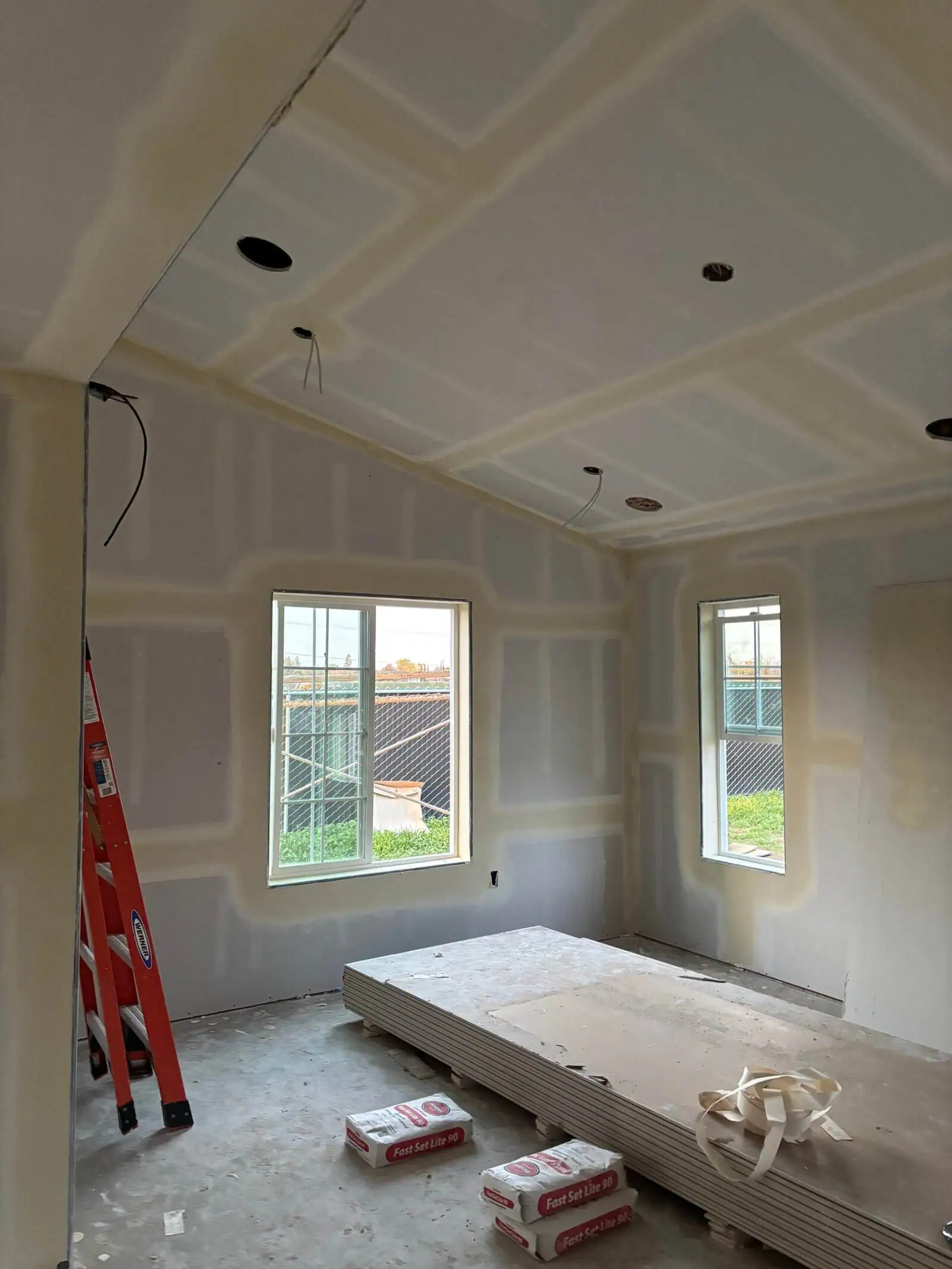 Drywall installation in a new residential room with windows and ceiling cutouts.